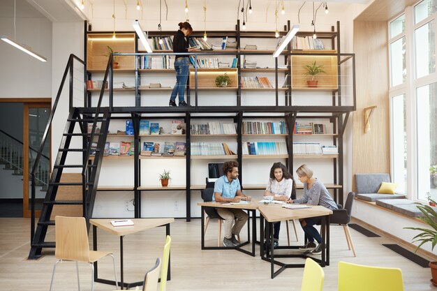 Three happy enthusiastic designers discussing buisness ideas for upcoming project sitting at table with papers in bright modern library. Comfort teamwork with friends. Startup, business concept