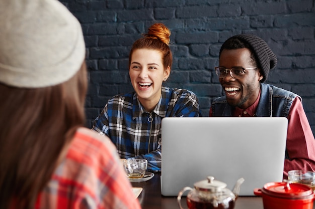 Три счастливых восторженных молодых людей с помощью портативного компьютера, в чате за столом в кафе. Международная команда обсуждает бизнес-идеи во время обеда.