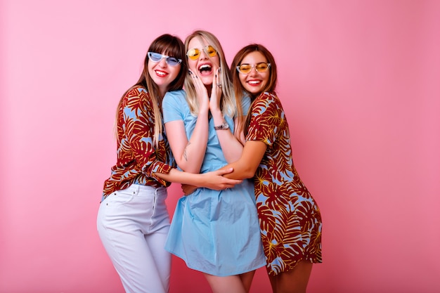 Три счастливые молодые красивые женщины, делающие селфи, веселая группа лучших друзей, стильная модная одежда с тропическим принтом и винтажные очки, розовая стена.