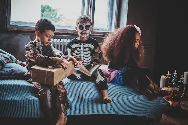 Three kids in Halloween costumes are sitting on the bed near window.
