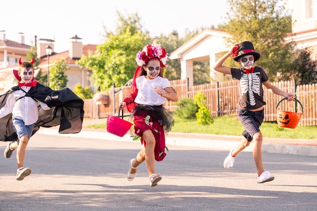 Three laughing kids in halloween costumes running down wide road on sunny autumn day while holding containers with sweet treats