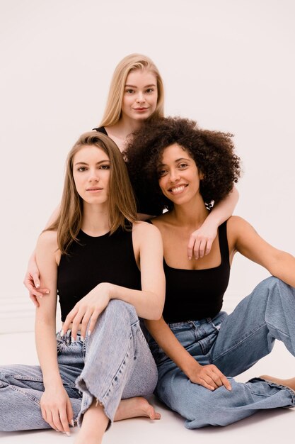 Три симпатичные молодые межрасовые женщины со светлыми и темными волосами в черных топах и джинсах смотрят в камеру на белом фоне