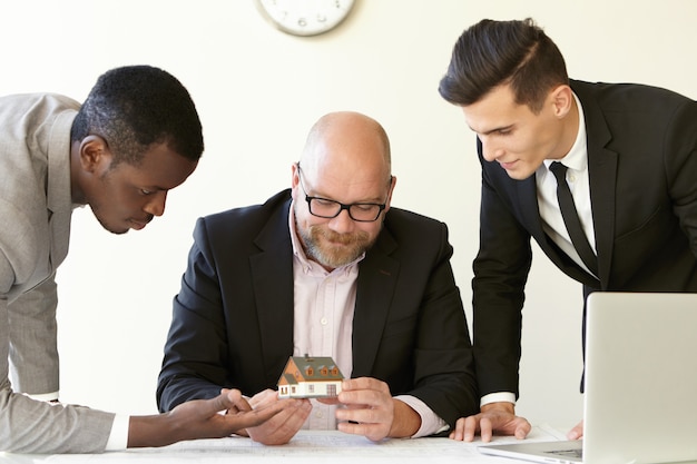 Free photo three office men estimating mock-up model of future terraced house. caucasian engineer in glasses holding miniature and smiling. other colleagues in suits looking at tiny house with interest.