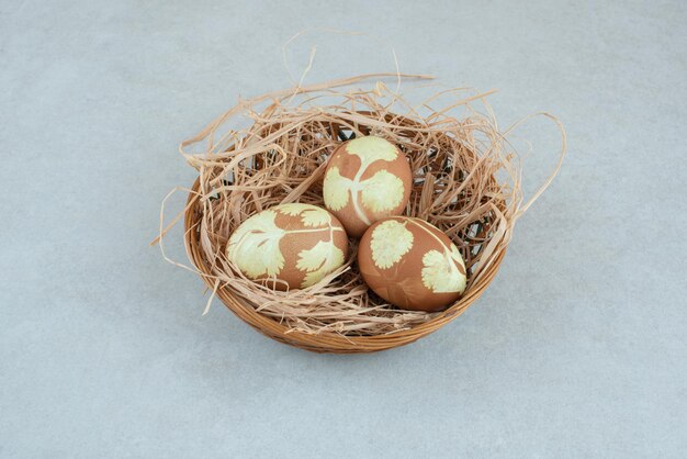 Three painted chicken eggs in hay on wicker basket .