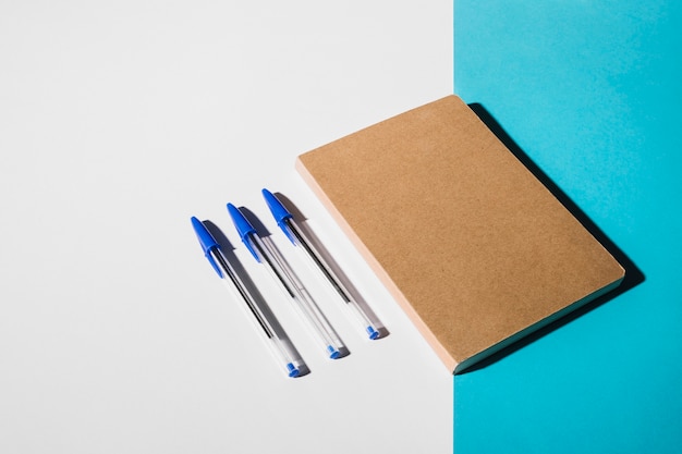 Three pens and closed book on dual white and blue backdrop