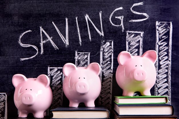 Three pink piggy banks standing on books next to a blackboard with savings chart.  