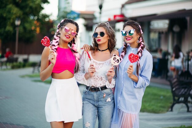 Three pretty and fashionable girls wearing in trendy clothes with colorful braid and professional makeup, after shopping. Women walking together, holding candies heart on stick and smiling.