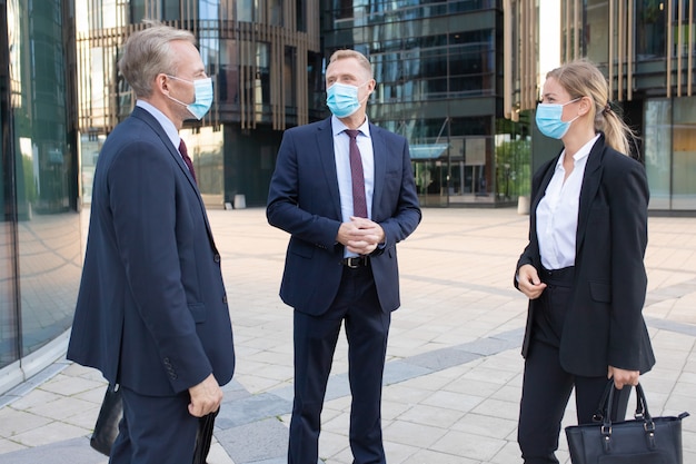外で取り引きを論議する顔のマスクの3人の専門のビジネス同僚。成功したマネージャーが通りに立って仕事について話しています。交渉、保護、パートナーシップの概念