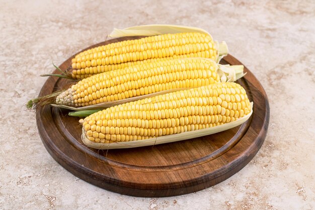 Three raw maize on wooden plate on marble surface