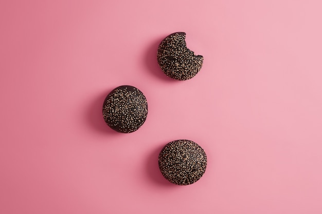 Free photo three round crusty charocal black burger buns with sesame seeds on rosy background. cuttlefish ink brioches for making sandwich, one is bitten. fast food and nutrition concept. view from above