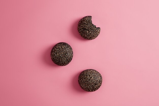 Three round crusty charocal black burger buns with sesame seeds on rosy background. Cuttlefish ink brioches for making sandwich, one is bitten. Fast food and nutrition concept. View from above