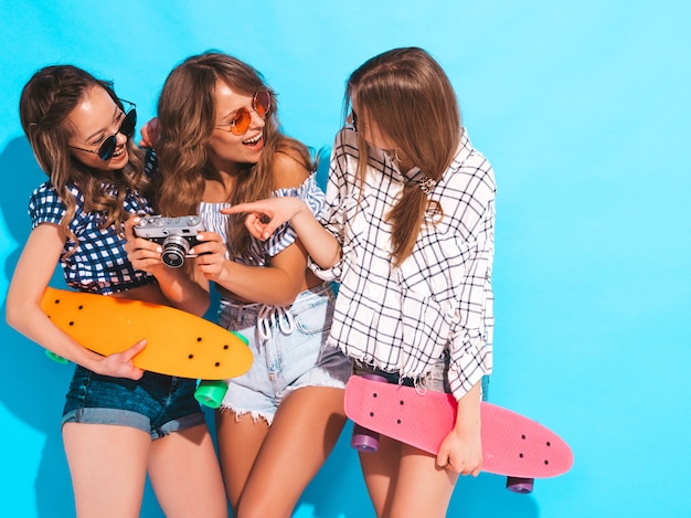 Three sexy beautiful stylish smiling girls with colorful penny skateboards. Women in summer checkered shirt clothes posing. Models taking pictures on retro photo camera