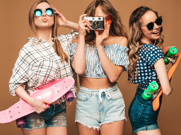 Three sexy beautiful stylish smiling girls with colorful penny skateboards. Women in summer checkered shirt clothes posing. Models taking pictures on retro photo camera