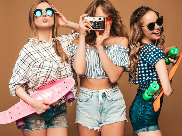 Three sexy beautiful stylish smiling girls with colorful penny skateboards. Women in summer checkered shirt clothes posing. Models taking pictures on retro photo camera