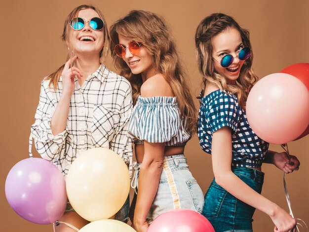 Три улыбающиеся красивые женщины в клетчатой рубашке летней одежды. Девочки позируют. Модели с разноцветными шарами в солнцезащитные очки. Веселимся, готовимся к празднованию дня рождения
