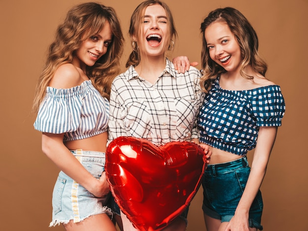 Three smiling beautiful women in checkered shirt summer clothes. Girls posing. Models with red heart shape balloon in sunglasses. Ready for celebration Valentine's Day