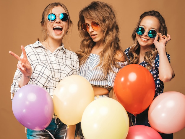 Три улыбающиеся красивые женщины в клетчатой рубашке летней одежды. Модели с разноцветными шарами в солнцезащитные очки. Веселимся, готовимся к празднованию дня рождения