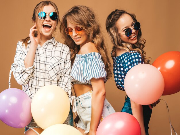 Three smiling beautiful women in checkered shirt summer clothes and sunglasses. Girls posing. Models with colorful balloons. Having fun, ready for celebration birthday party