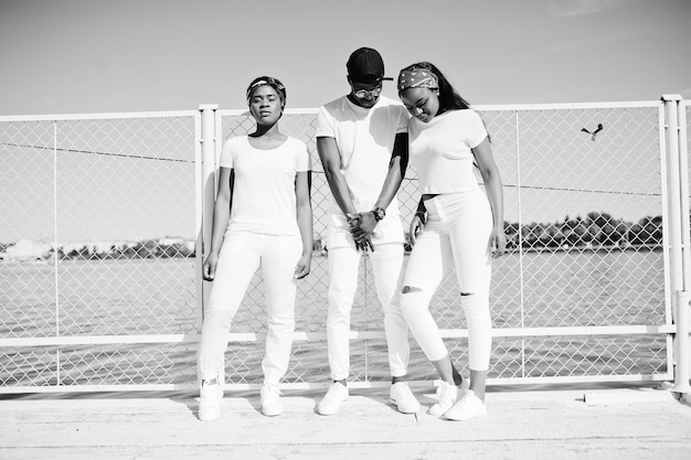 Three stylish african american friends wear on white clothes at pier on beach against cage Street fashion of young black people Black man with two african girls