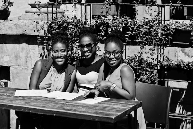 晴れた夏の日の屋外でポーズをとった3人のスタイリッシュなアフリカ系アメリカ人女性が手元にメニューを持つレストランのテーブルに座っています。