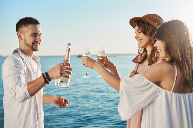 Foto gratuita tre giovani attraenti e alla moda che si levano in piedi sul mare e bevono mentre sorridono ampiamente, parlando di qualcosa. i colleghi trascorrono il tempo libero alla festa organizzata dall'azienda.