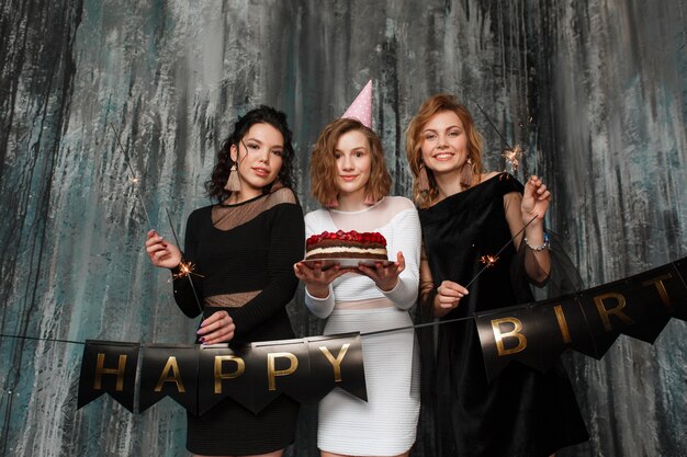 Three young beautiful girls celebrate birthday holding cake and sparklers 