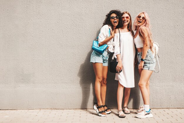 Three young beautiful smiling female in trendy summer clothesSexy carefree multiracial women posing on the street background near wallPositive models having fun in sunglasses Cheerful and happy