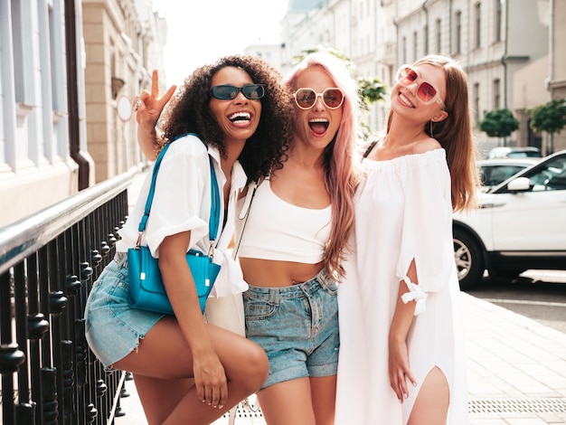 트렌디한 여름 옷을 입은 3명의 젊고 아름다운 미소 거리 배경에서 포즈를 취하는 섹시하고 평온한 다인종 여성선글라스를 쓴 긍정적인 모델 명랑하고 행복한