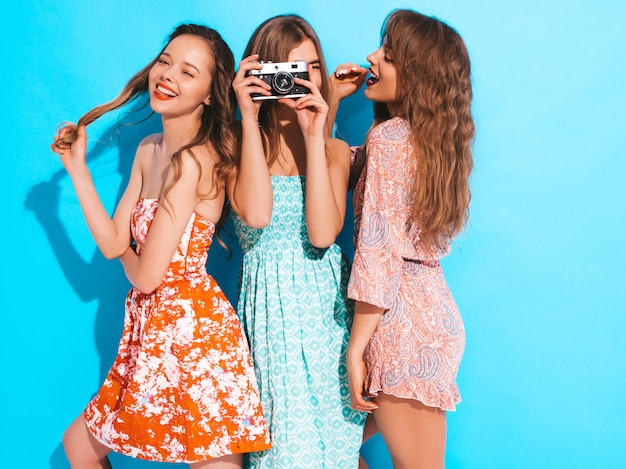 トレンディな夏のカジュアルドレスの3人の若い美しい笑顔の女の子。セクシーな屈託のない女性がポーズします。レトロなカメラで写真を撮る