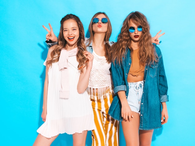 Three young beautiful smiling girls in trendy summer colorful clothes. Sexy carefree women in sunglasses isolated on blue. Positive models