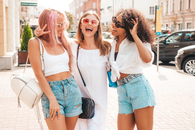 Три молодые красивые улыбающиеся женщины-хипстеры в модной летней одеждеСексуальные беззаботные многорасовые женщины позируют на фоне улицыПозитивные модели веселятся в солнцезащитных очках Веселые и счастливые