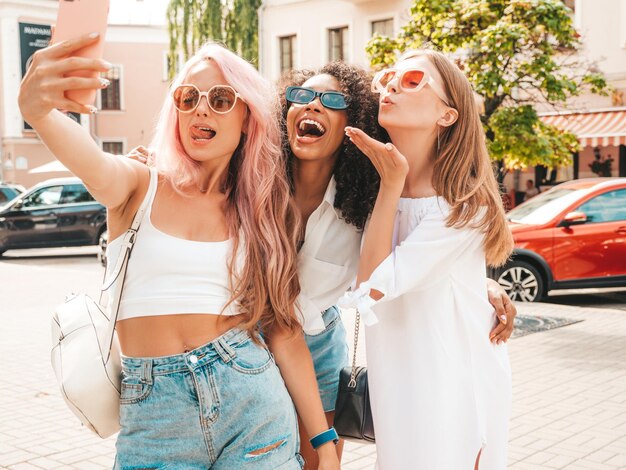 트렌디한 여름 옷을 입은 3명의 젊고 아름다운 힙스터 여성거리 배경에서 포즈를 취하는 섹시하고 평온한 다인종 여성선글라스를 착용하고 즐거운 시간을 보내는 긍정적인 모델 셀카 사진 찍기