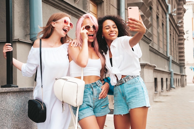 Три молодые красивые улыбающиеся женщины-хипстеры в модной летней одеждеСексуальные беззаботные многорасовые женщины позируют на фоне улицыПозитивные модели веселятся в солнцезащитных очках Делая селфи-фотографии