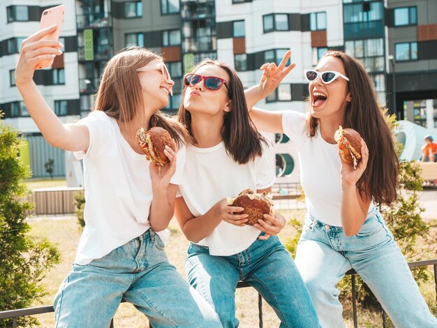 トレンディな夏の服を着た3人の若い美しい笑顔のヒップスター女性ストリートでポーズをとるセクシーな屈託のない女性サングラスで自分撮りをするポジティブモデルジューシーなハンバーガーを持ってハンバーガーを食べる