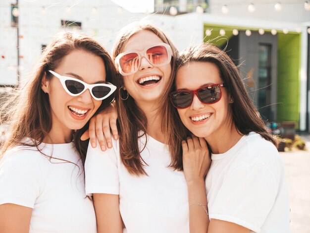 Three young beautiful smiling hipster female in trendy summer same clothes Sexy carefree women posing on the street backgroundPositive models having fun in sunglassesHugging Cheerful and happy