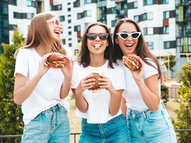 Three young beautiful smiling hipster female in trendy summer same clothes Sexy carefree women posing in the streetPositive models having fun in sunglassesHolding juicy burger and eating hamburger