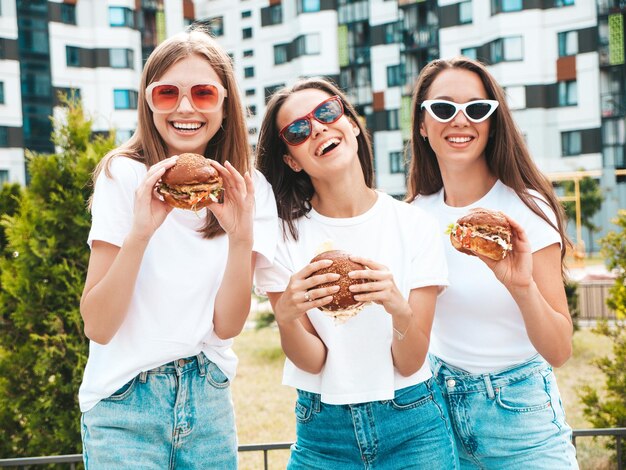 トレンディな夏の同じ服を着た3人の若い美しい笑顔のヒップスター女性ストリートでポーズをとるセクシーな屈託のない女性サングラスで楽しんでいるポジティブモデルジューシーなハンバーガーを持ってハンバーガーを食べる