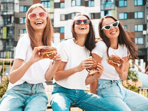 トレンディな夏の同じ服を着た3人の若い美しい笑顔のヒップスター女性ストリートでポーズをとるセクシーな屈託のない女性サングラスで楽しんでいるポジティブモデルジューシーなハンバーガーを持ってハンバーガーを食べる