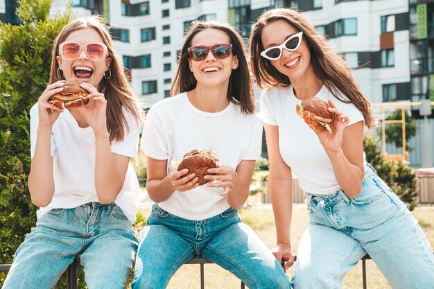 トレンディな夏の同じ服を着た3人の若い美しい笑顔のヒップスター女性ストリートでポーズをとるセクシーな屈託のない女性サングラスで楽しんでいるポジティブモデルジューシーなハンバーガーを持ってハンバーガーを食べる