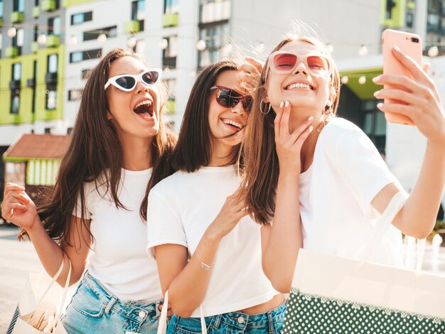 Three young beautiful smiling hipster female in trendy summer same clothes Sexy carefree women posing in the streetPositive models having fun in sunglassesHolding shopping bagsTaking selfie photos