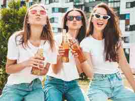 Free photo three young beautiful smiling hipster female in trendy summer same clothessexy carefree women posing in the streetpositive models having fun in sunglasses drinking bottle beer oktoberfest