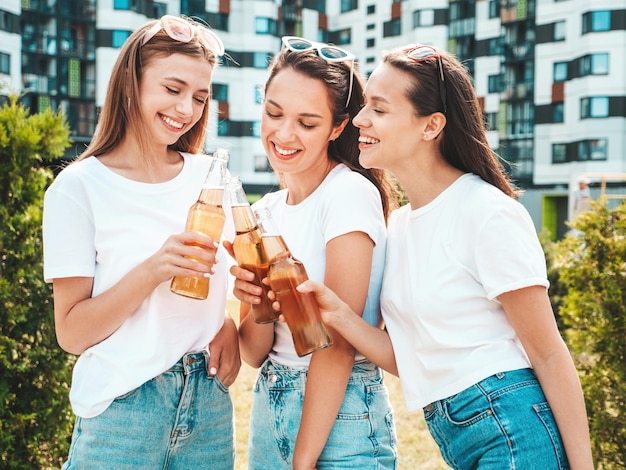 Free photo three young beautiful smiling hipster female in trendy summer same clothessexy carefree women posing in the streetpositive models having fun in sunglasses drinking bottle beer oktoberfest
