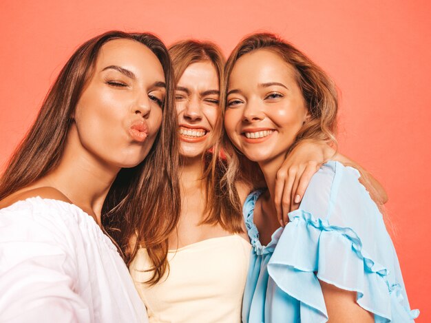 Three young beautiful smiling hipster girls in trendy summer clothes. Sexy carefree women posing near pink wall. Positive models going crazy.Taking selfie self portrait photos on smartphone