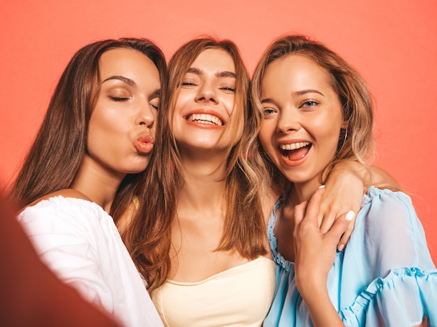 トレンディな夏服で3人の若い美しい笑顔流行に敏感な女の子。ピンクの壁に近いポーズセクシーな屈託のない女性。ポジティブなモデルが夢中になります。スマートフォンでセルフポートレート写真を撮る