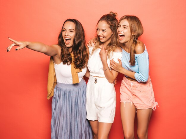 최신 유행의 여름 옷을 입고 세 젊은 아름 다운 웃는 hipster 여자. 분홍색 벽 근처 포즈 섹시 평온한 여성. 긍정적 인 모델 재미. 상점 판매를 가리키는