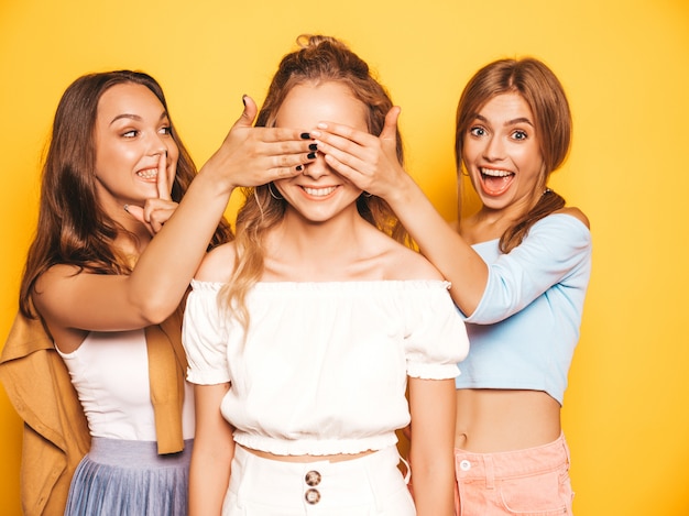 Three young beautiful smiling hipster girls in trendy summer clothes.Sexy carefree women posing near yellow wall.Models surprising their friend.They cover her eyes and hugging from behind