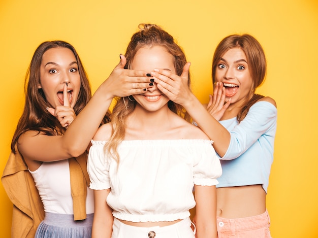 トレンディな夏服の3人の若い美しい笑顔流行に敏感な女の子。黄色の壁の近くでポーズをとってセクシーな屈託のない女性。彼らの友人を驚かせるモデル。