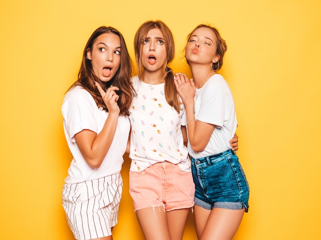 Three young beautiful smiling hipster girls in trendy summer clothes. Sexy carefree women posing near yellow wall. Positive models going crazy and having fun.Hugging