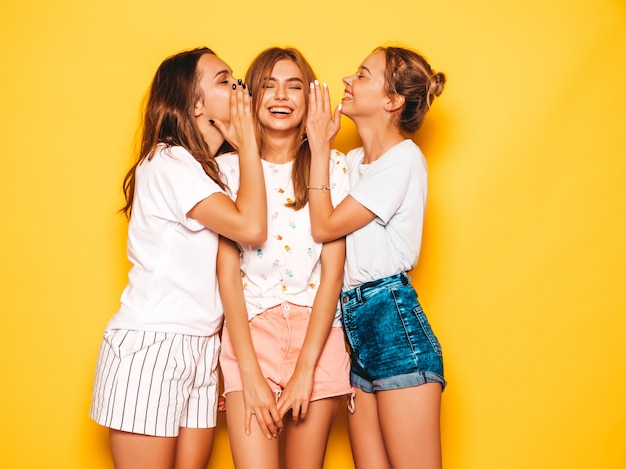トレンディな夏服で3人の若い美しい笑顔流行に敏感な女の子。黄色の壁に近いポーズセクシーな屈託のない女性。夢中になって楽しんでいるポジティブなモデル。秘密を共有し、ゴシップ