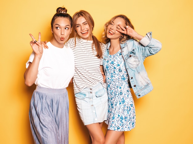 Three young beautiful smiling hipster girls in trendy summer clothes. Sexy carefree women posing near yellow wall. Positive models having fun